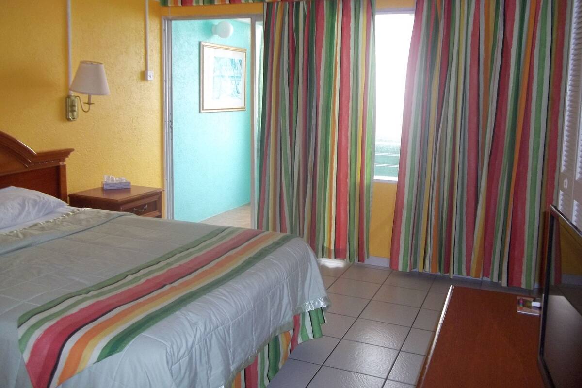 Une chambre d’hôtel avec un lit, une table de chevet, une lampe, une porte avec des rideaux à rayures et une vue à travers la porte.