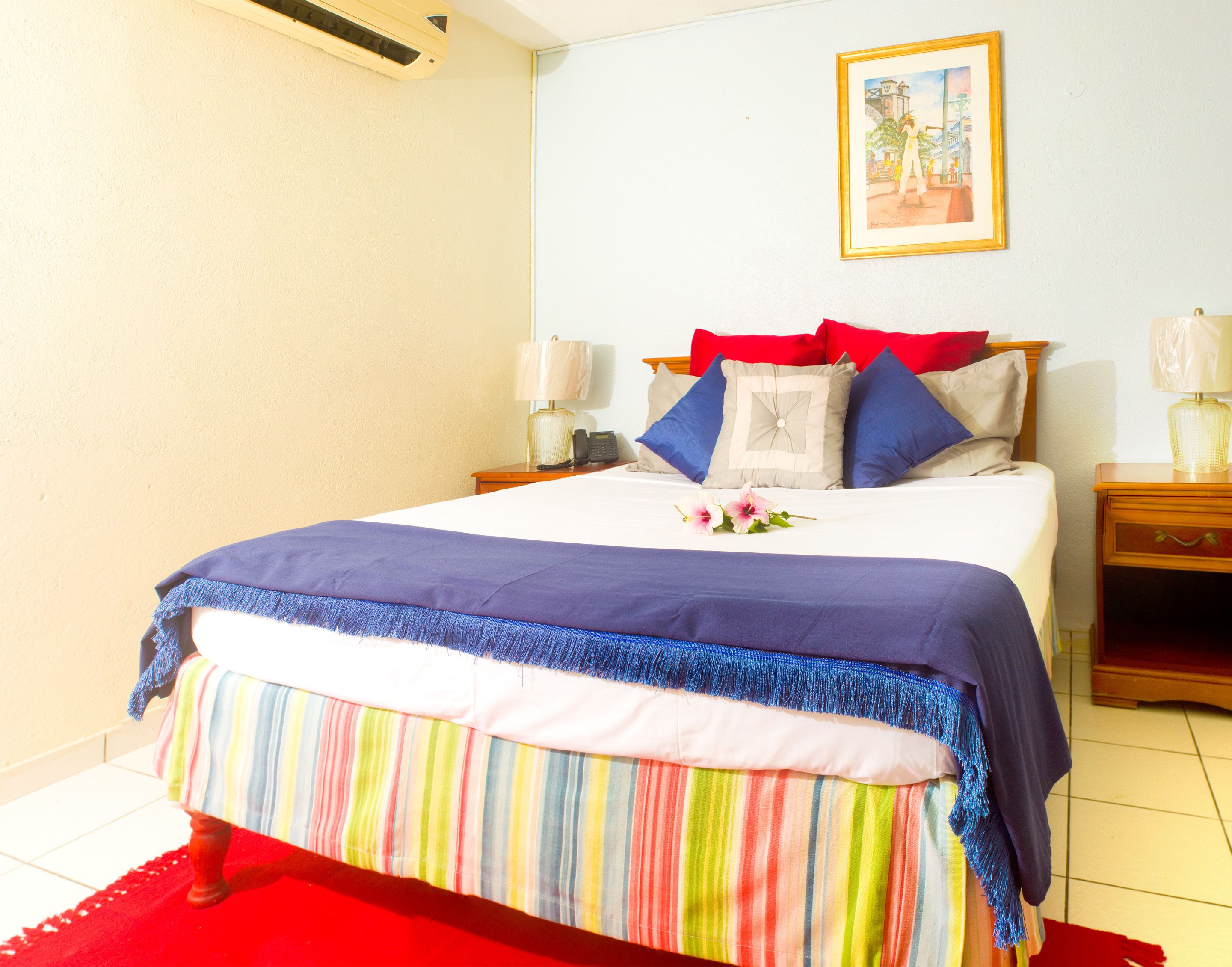 Un lit bien fait, agrémenté d’une jupe de lit rayée et colorée, d’une couvre-lit rouge et bleue, et d’une seule fleur en guise de décoration. Un tableau encadré orne le mur, une table de chevet avec une lampe s’y trouve, ainsi qu’une petite table de nuit également équipée d’une lampe.