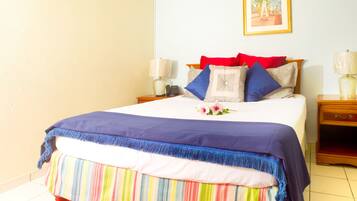 Una cama bien tendida con falda a rayas de colores, colcha azul y roja, y una sola flor decorativa. En la pared, un cuadro enmarcado; una mesita de noche con lámpara, y un tocador con lámpara.