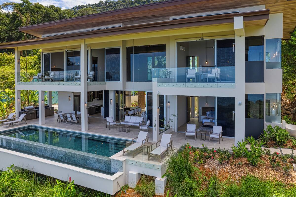 A modern multi-story house with a large pool, outdoor seating, and a lush garden.