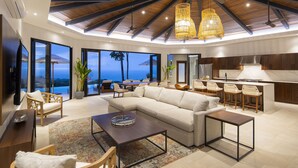 Un salón amplio con un sofá grande, una mesa de centro y un comedor con sillas. Cuenta con una cocina de madera y un ventanal con vistas al océano.