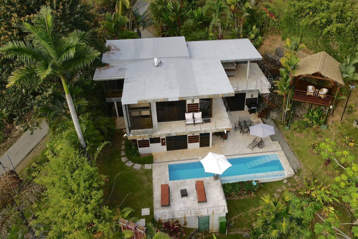 A two-story house with a swimming pool, surrounded by lush greenery and palm trees.