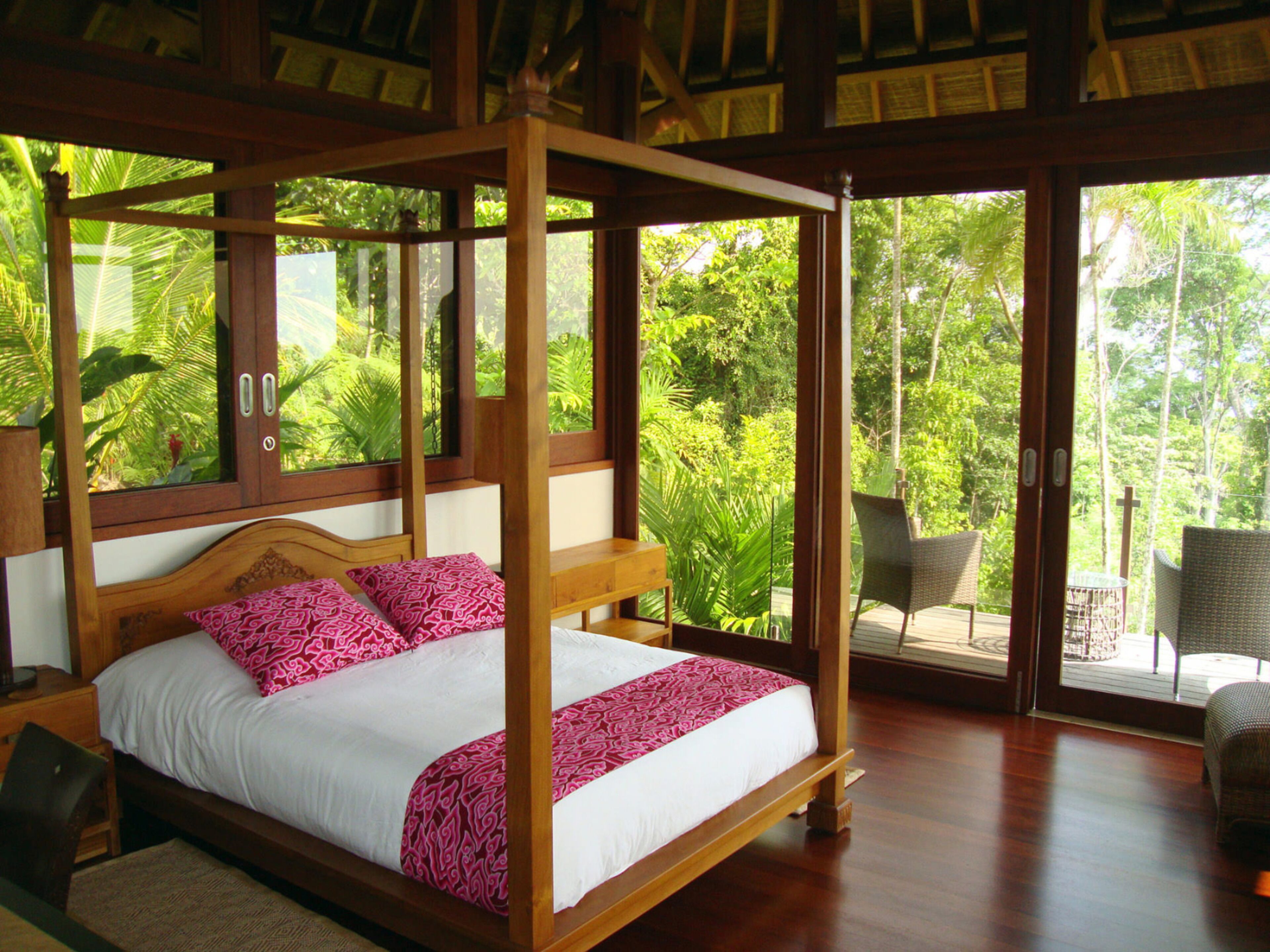 Una cama con dosel de madera y ropa de cama blanca, ubicada en una habitación con ventanas amplias que dan a un exuberante follaje.