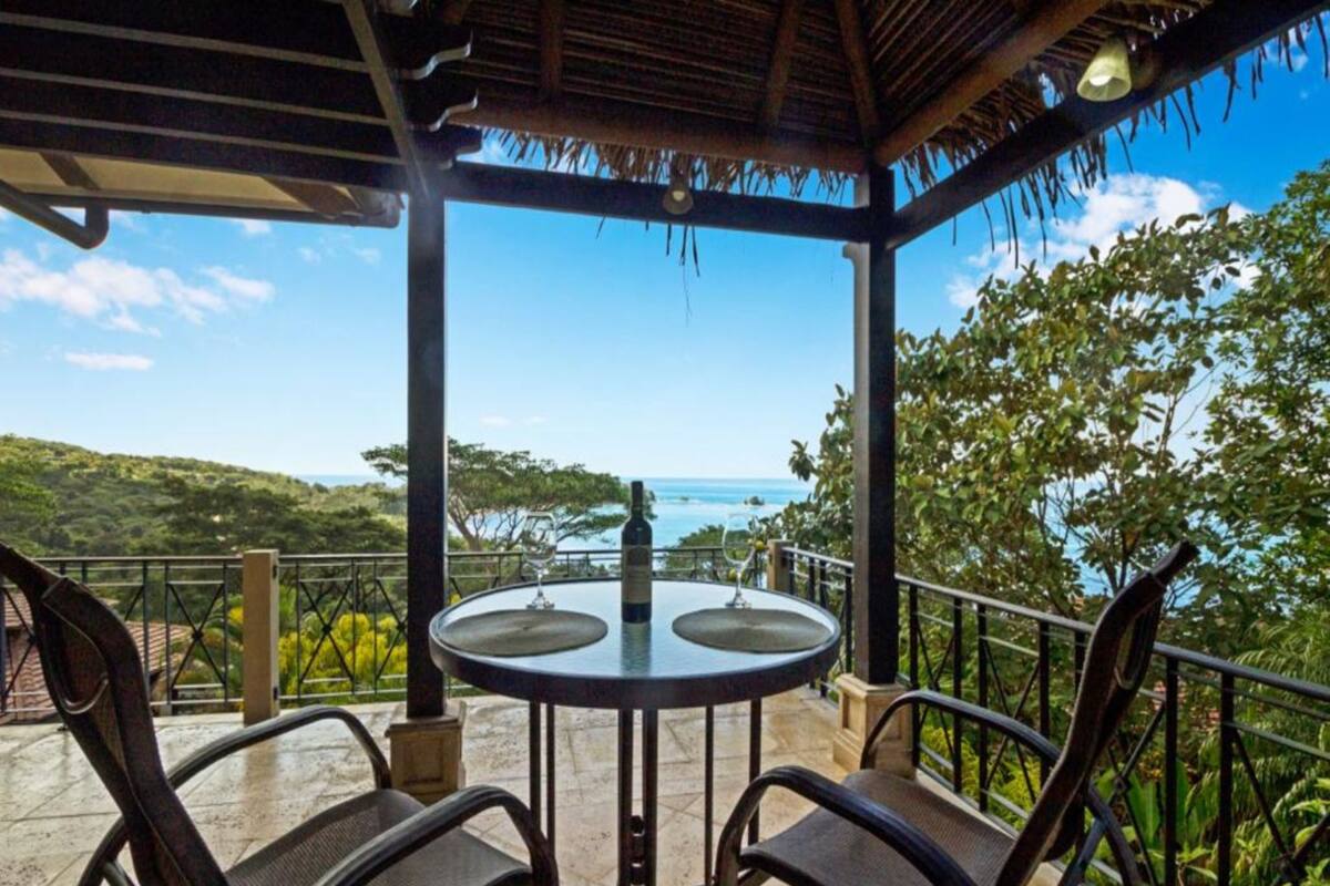 A covered outdoor seating area with a table and chairs, overlooking a scenic view of the ocean and hills.