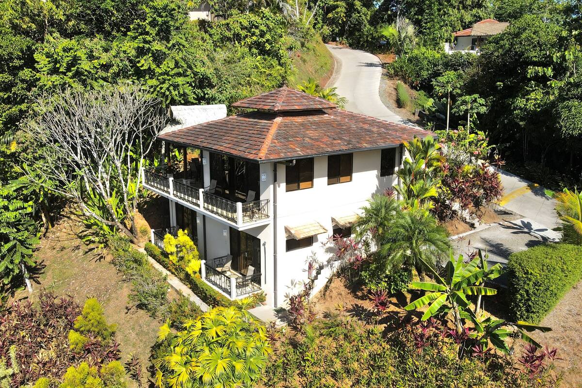 A two-story house with a balcony, surrounded by lush greenery and a curved road.