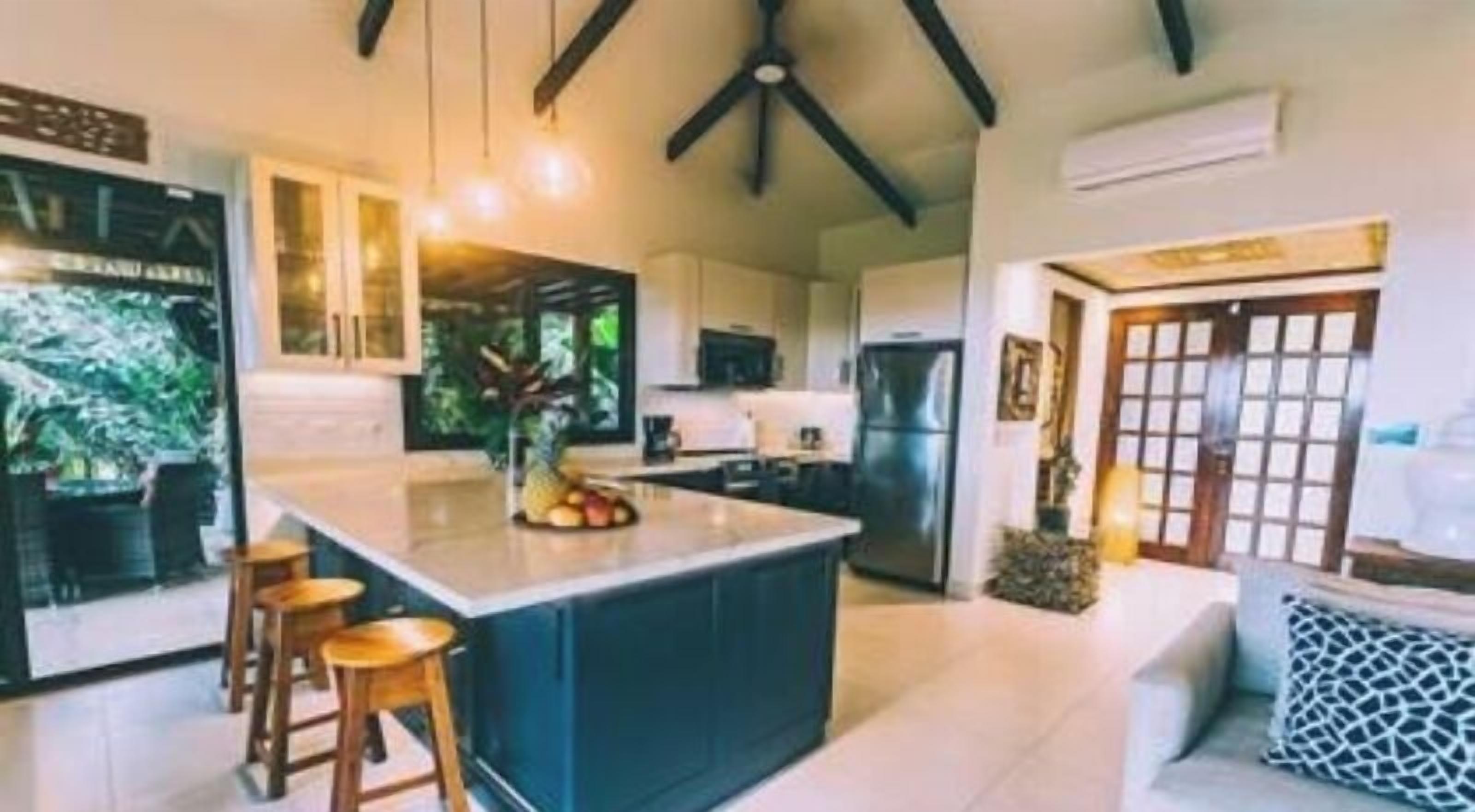 A modern kitchen with a central island, bar stools, and a dining area with a sofa and patterned cushions.