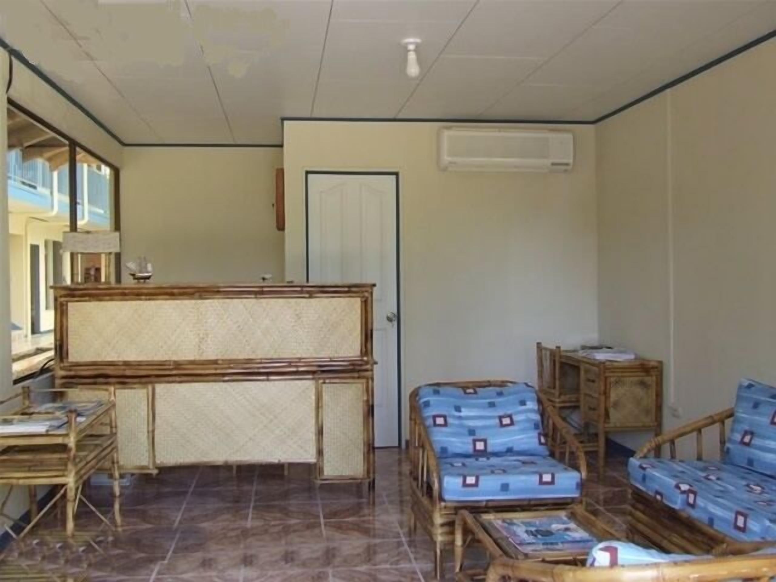 A room with a wooden reception desk, two wicker chairs, a sofa, and a tiled floor.
