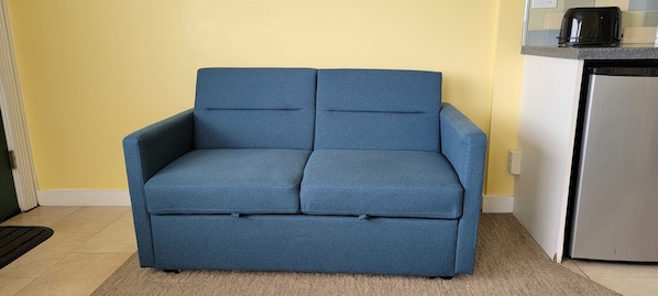 A blue sofa in a room with yellow walls, a kitchen area, and a tiled floor. - Sea Sprite at Haystack Rock (Cannon Beach)