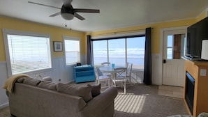 A living room with a sofa, a small table, and a chair. There is a large window with blinds, a ceiling fan, and a door leading outside. - Sea Sprite at Haystack Rock (Cannon Beach)