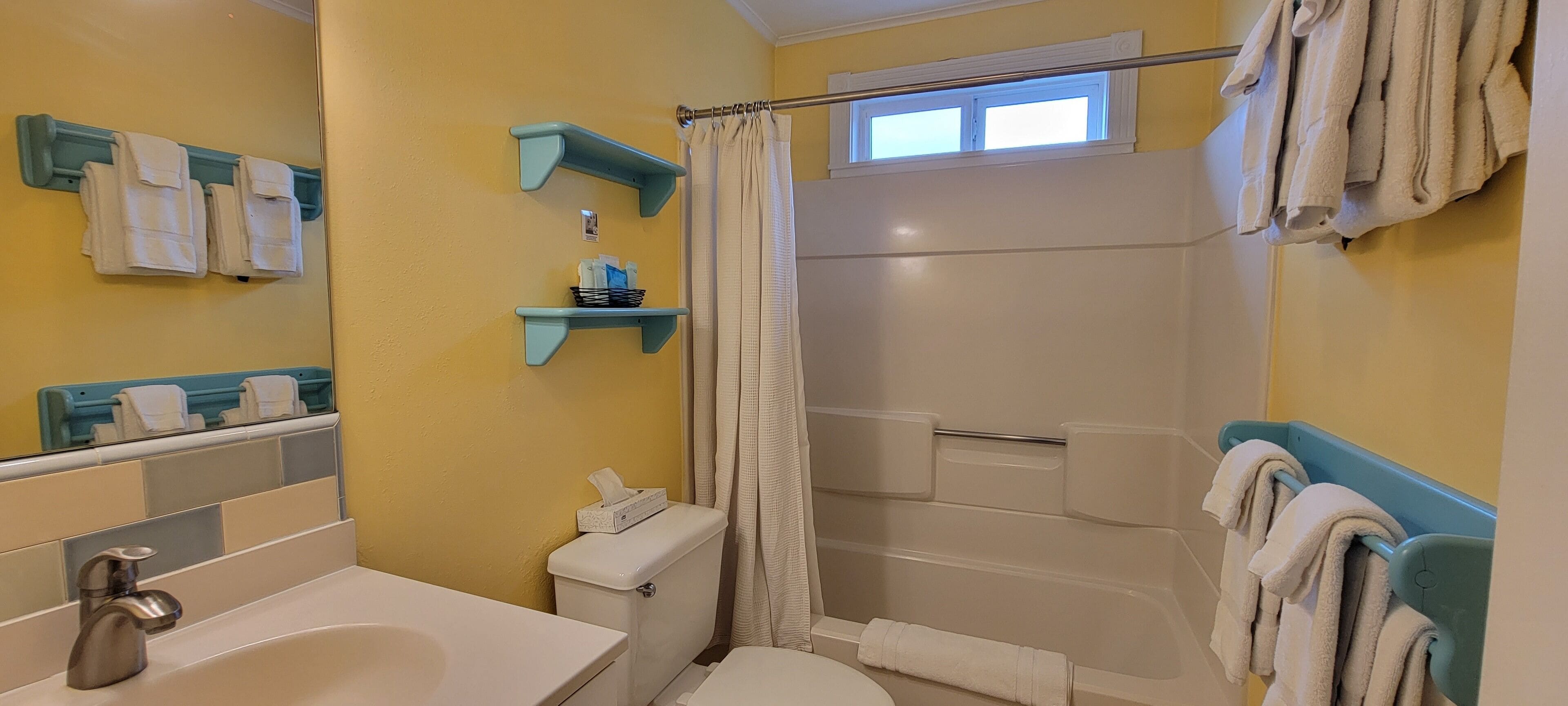 Uma casa de banho com banheira, sanita e lavatório. Há porta-toalhas e uma janela sobre o chuveiro.