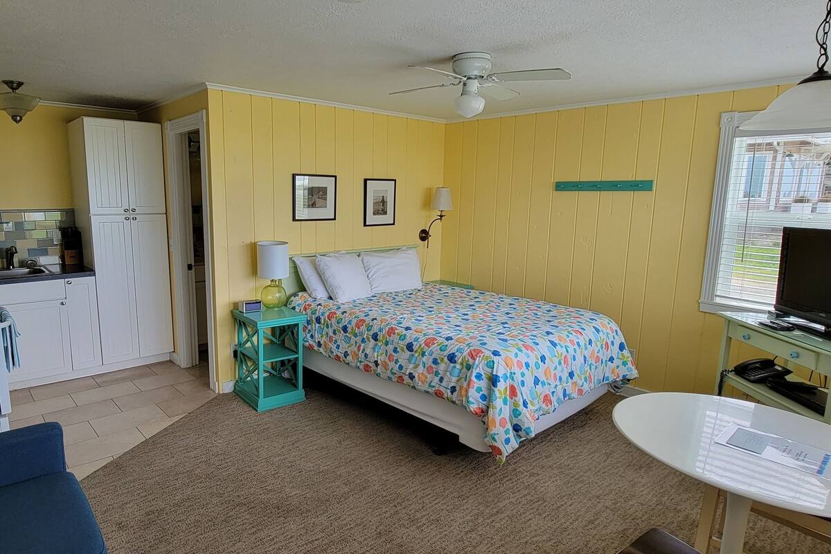 Une chambre d’hôtel compacte comprenant un coin cuisine, un lit recouvert d’une courtepointe à motifs floraux colorés, un ventilateur de plafond, une télévision et une petite table avec une chaise.