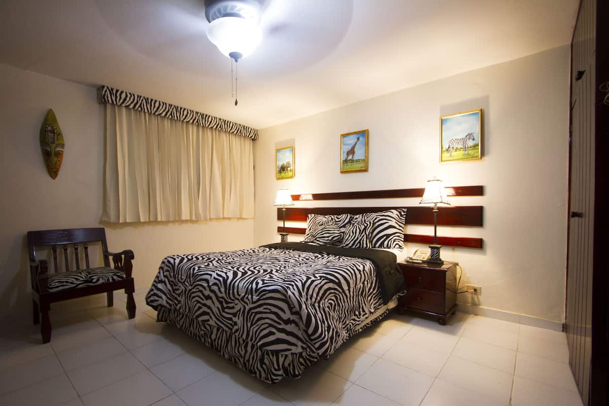 A bedroom with a zebra-patterned bedspread, a wooden bench, a nightstand with a lamp, and wall art depicting animals.