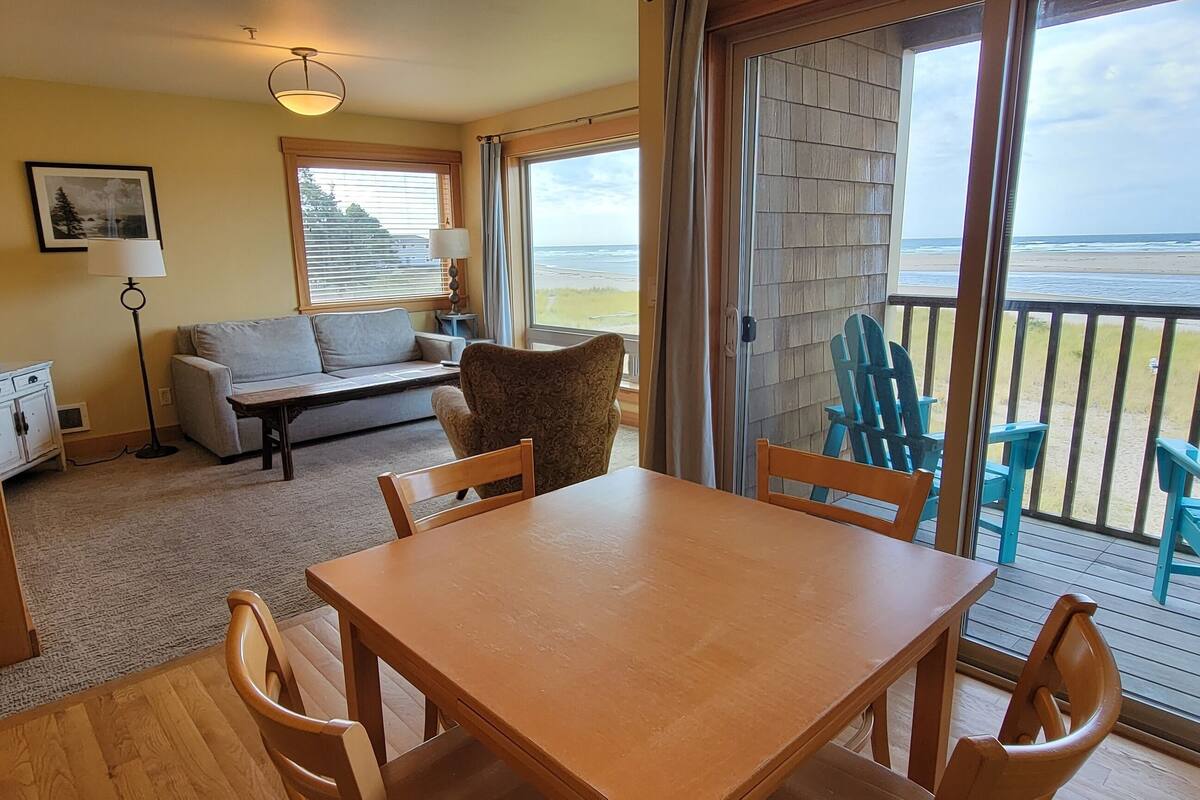 A room with a wooden table and chairs, a sofa, a lamp, and a view of the beach through large windows.