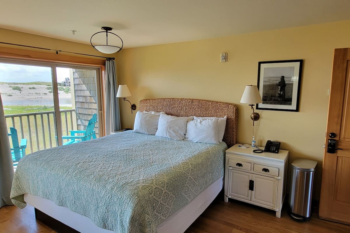 A bedroom with a bed, a nightstand, a lamp, a picture on the wall, a door, and a balcony with a chair.