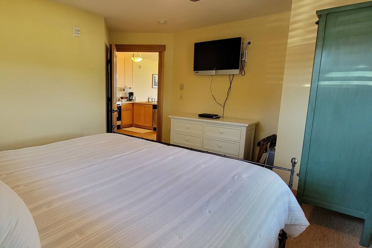 A hotel room with a bed, a dresser, a television, and a door leading to a kitchen area.