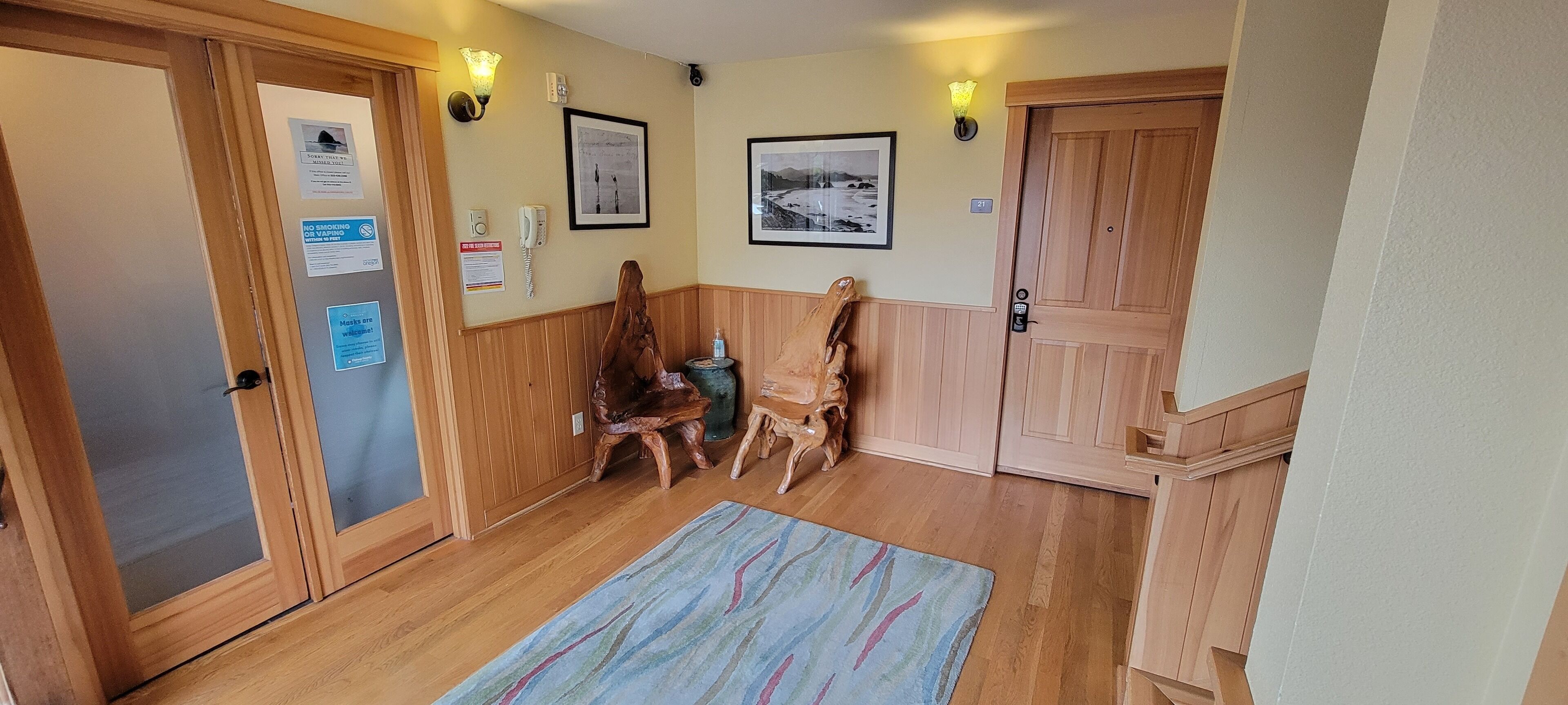 A hallway with wooden paneling, a wooden door, a wall-mounted phone, framed pictures, and a small table with a chair.