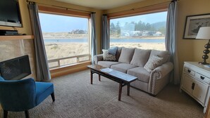A room with a sofa, a coffee table, a TV, and a view of a beach and houses. - Sea Sprite on the Estuary (Cannon Beach)
