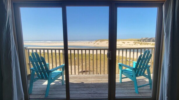 A view from a room with a sliding glass door to a balcony with two turquoise chairs and a wooden deck. - Sea Sprite on the Estuary (Cannon Beach)
