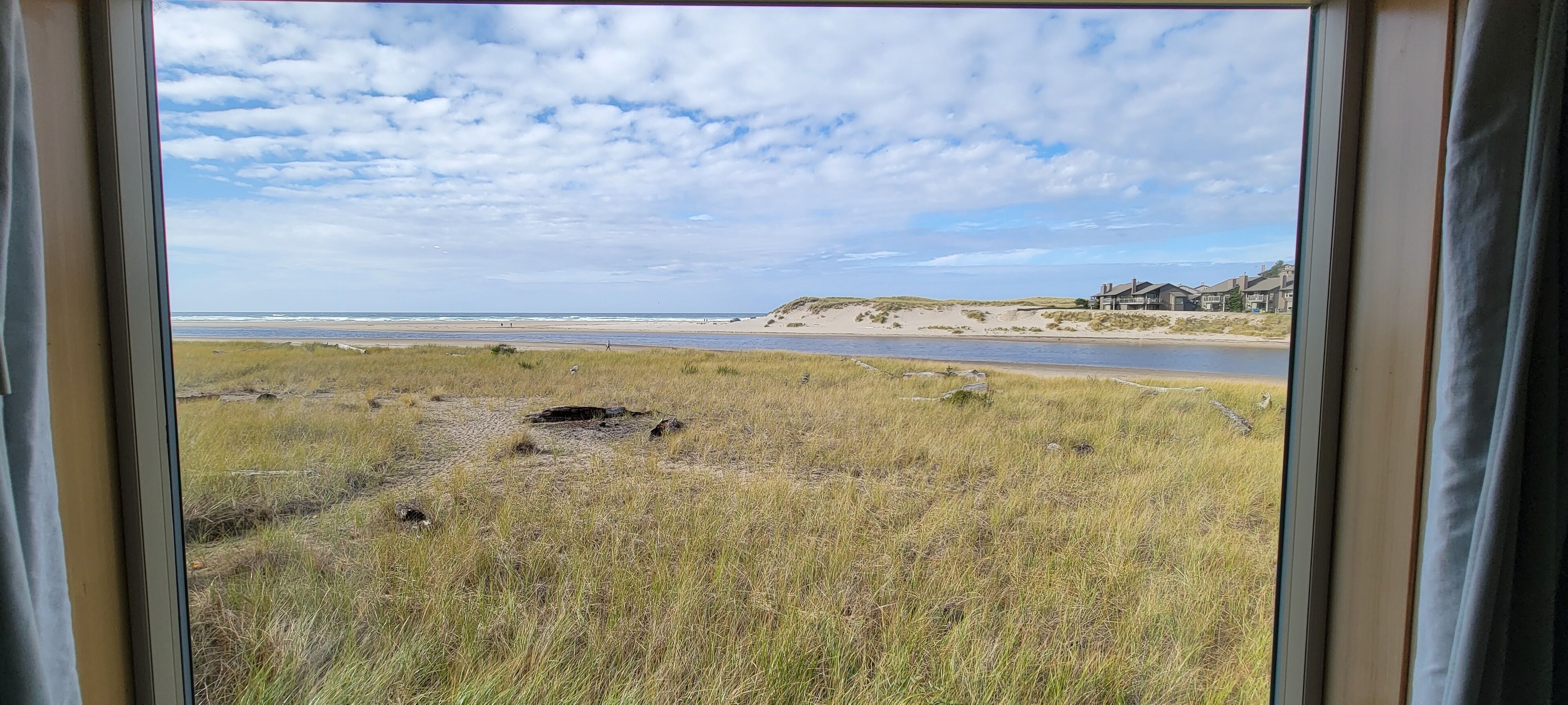 A view through a window of a beach with grassy dunes, a river, and a building in the distance.