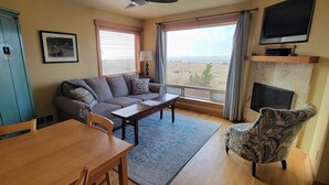 A living room with a sofa, a wooden table, a patterned armchair, a fireplace, and a TV mounted above it. - Sea Sprite on the Estuary (Cannon Beach)