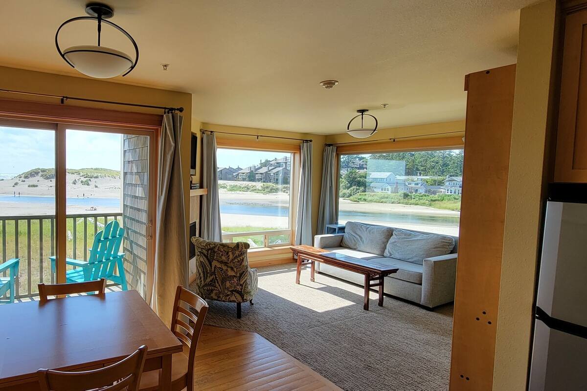 A room with a dining table, chairs, a sofa, and a view of the beach.