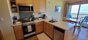 Cocina compacta con microondas, estufa y lavabo. Hay una zona de comedor con mesa y sillas, y vistas a la playa a través de grandes ventanales.