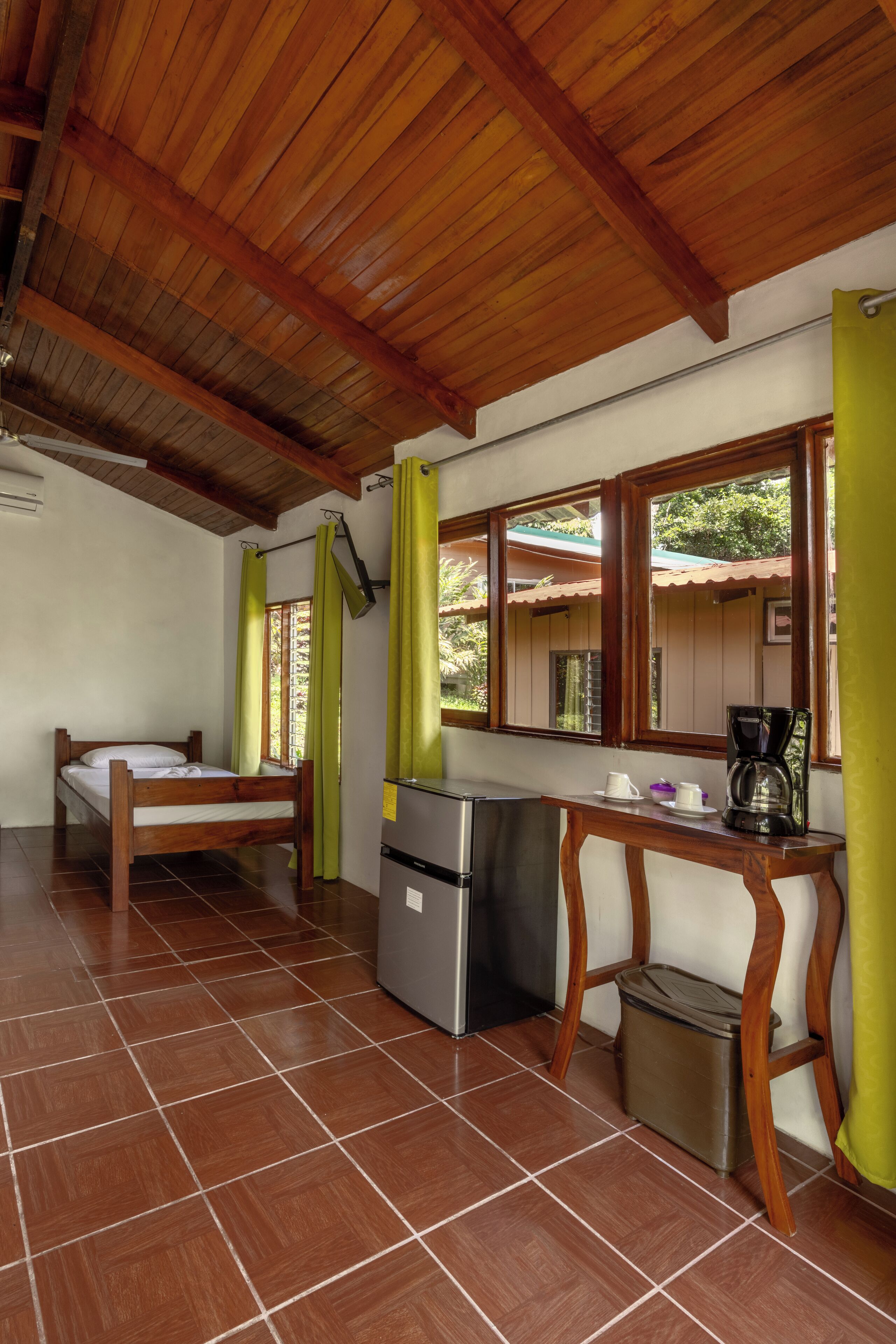 A room with a bed, a small table with a coffee maker, a trash bin, and a window with curtains.