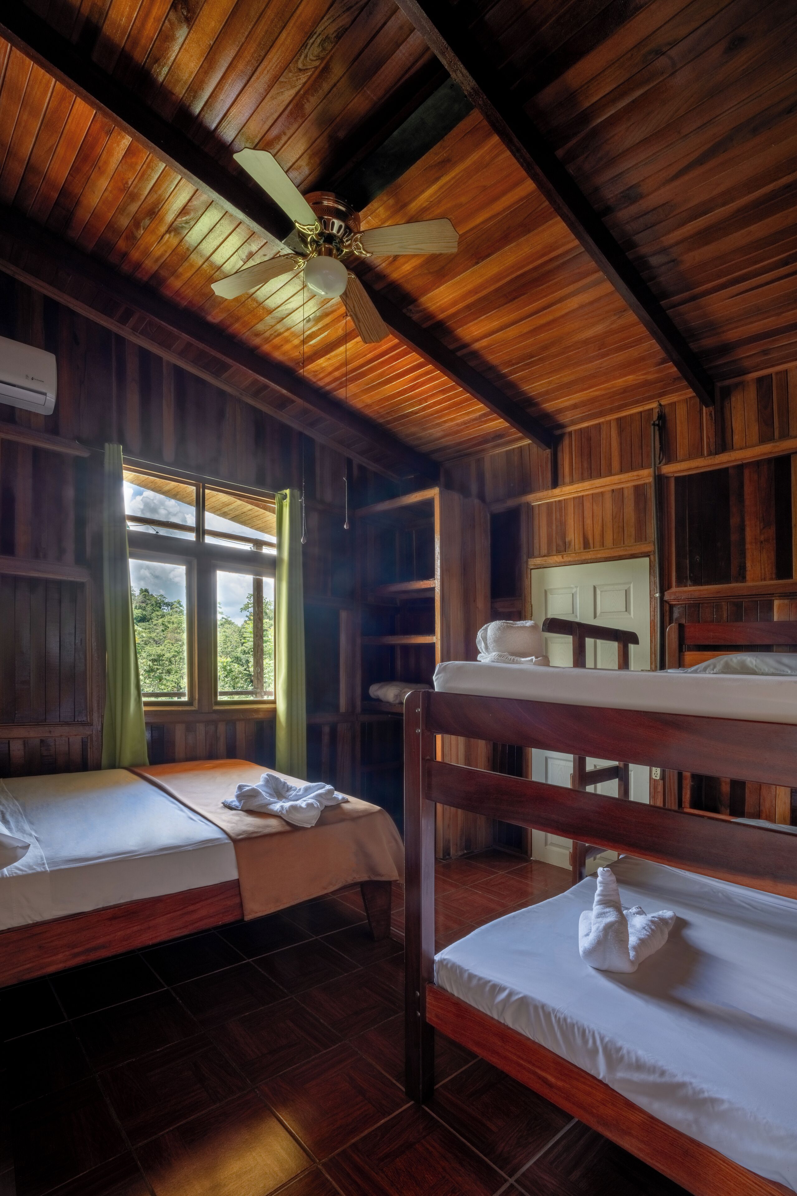 A wooden cabin room with two bunk beds, a ceiling fan, and a window with curtains.