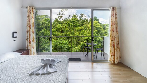 A bedroom with a large bed, a balcony with a view of lush greenery, a small table and chairs, and a wall-mounted phone. - Villas Josipek (Peñas Blancas)