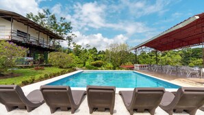A swimming pool with lounge chairs and a covered area, surrounded by greenery and a building. - Villas Josipek (Peñas Blancas)