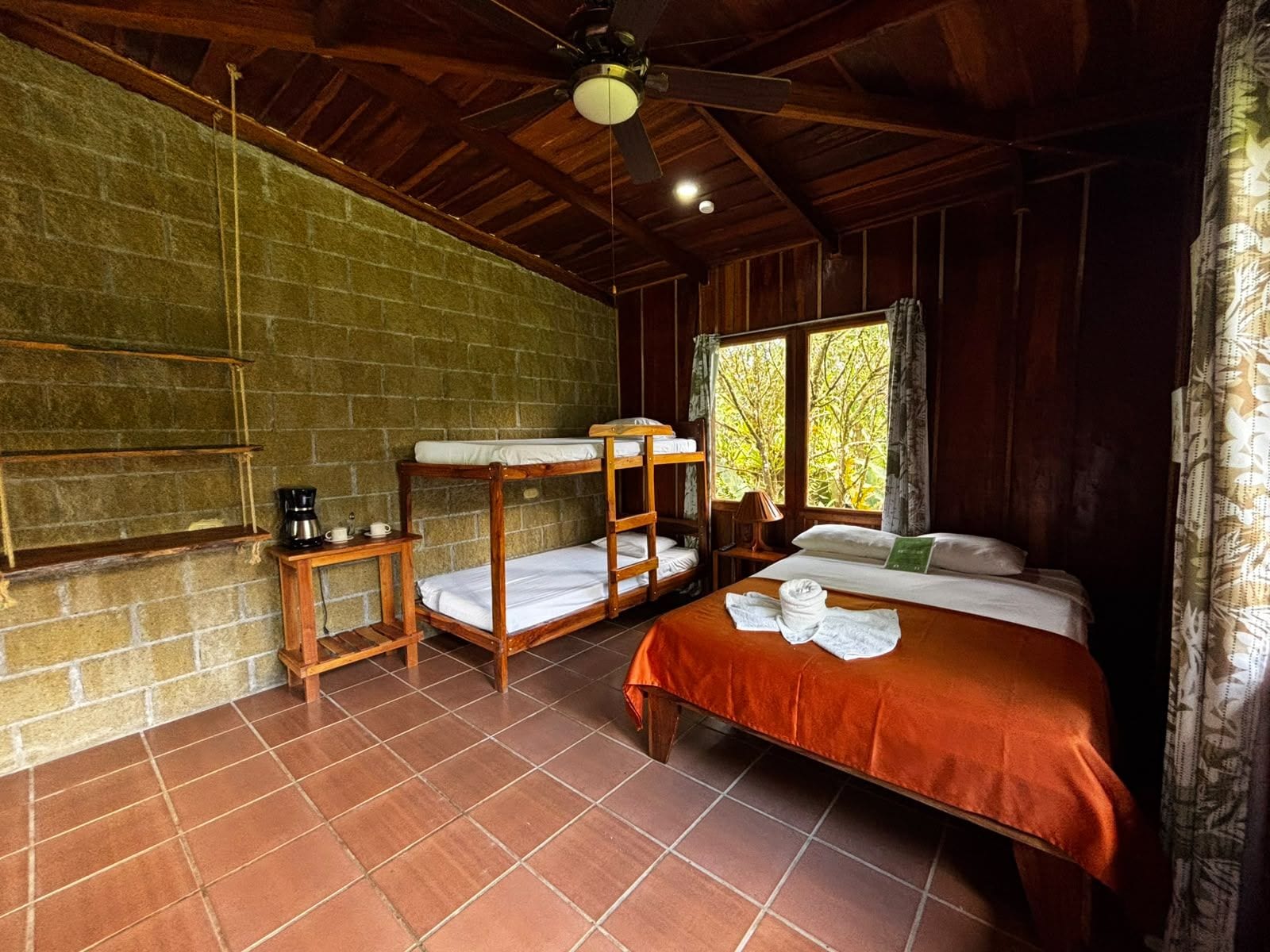 Habitación rústica con pared de ladrillo, camas literas de madera, una cama individual, una mesita con cafetera y ventana con vista a árboles.