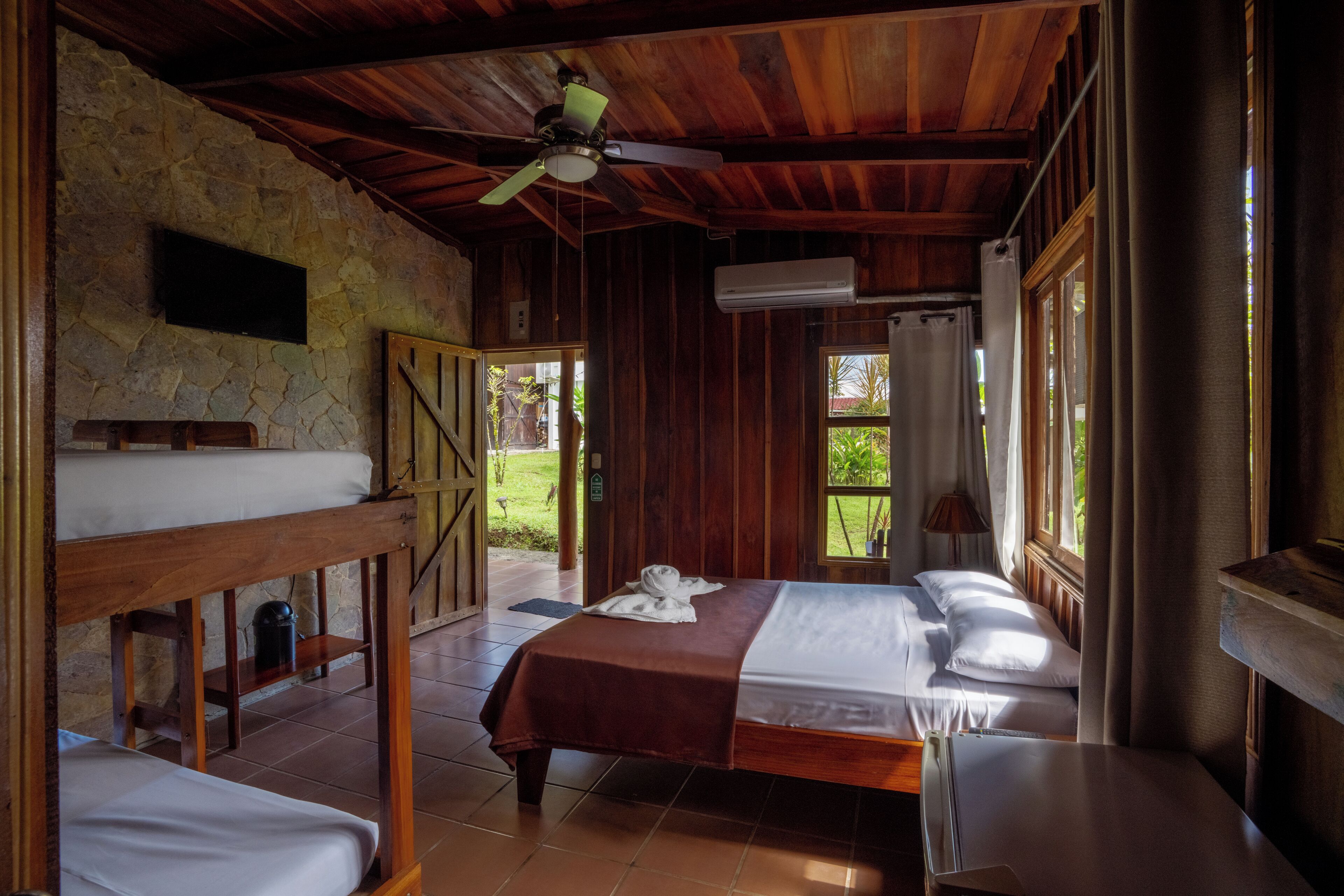 Una habitación rústica de madera con literas, un ventilador de techo, una pared de piedra y una ventana con cortinas.