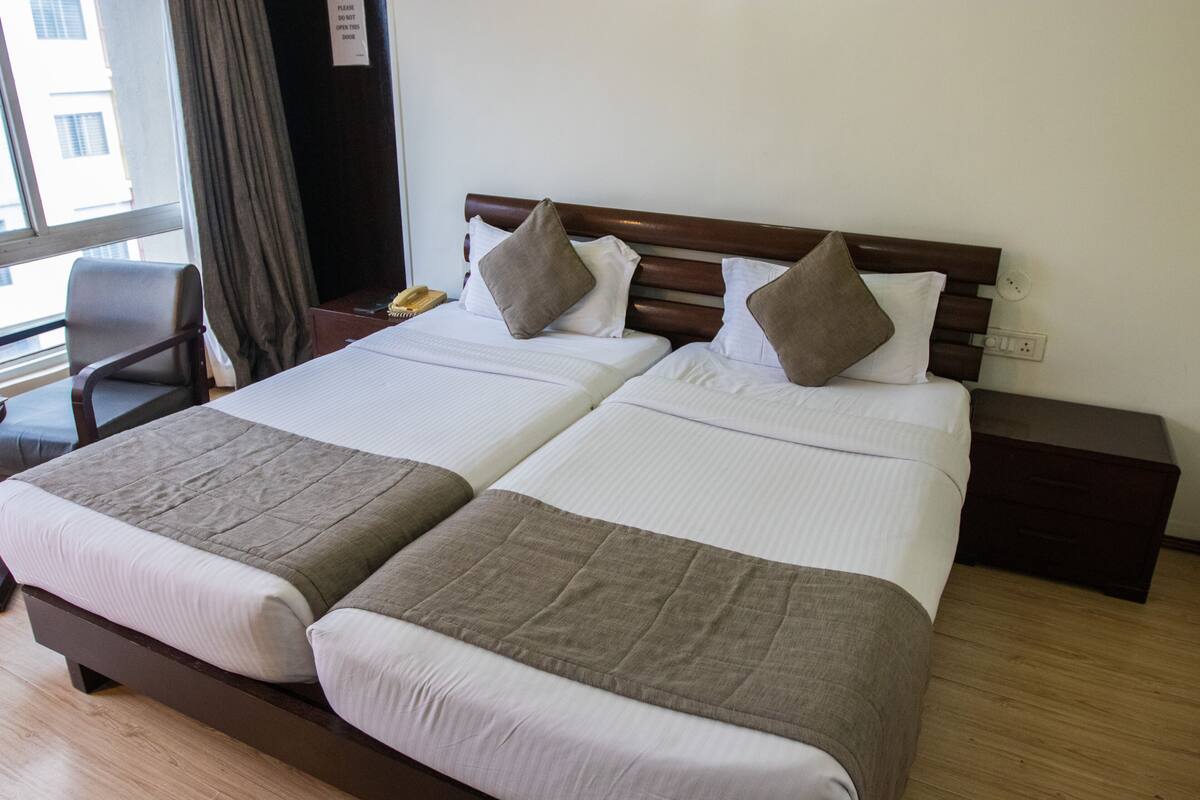 Habitación de hotel con dos camas individuales, cabecero de madera, mesita de noche con lámpara y una silla.
