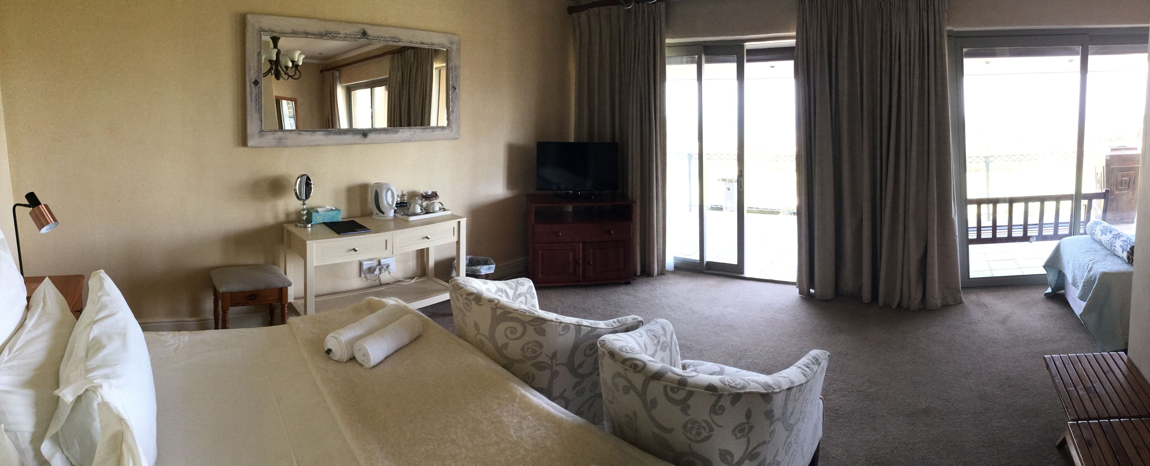 A hotel room with a bed, a desk with a mirror, a television, and a view of the outside through a sliding door.