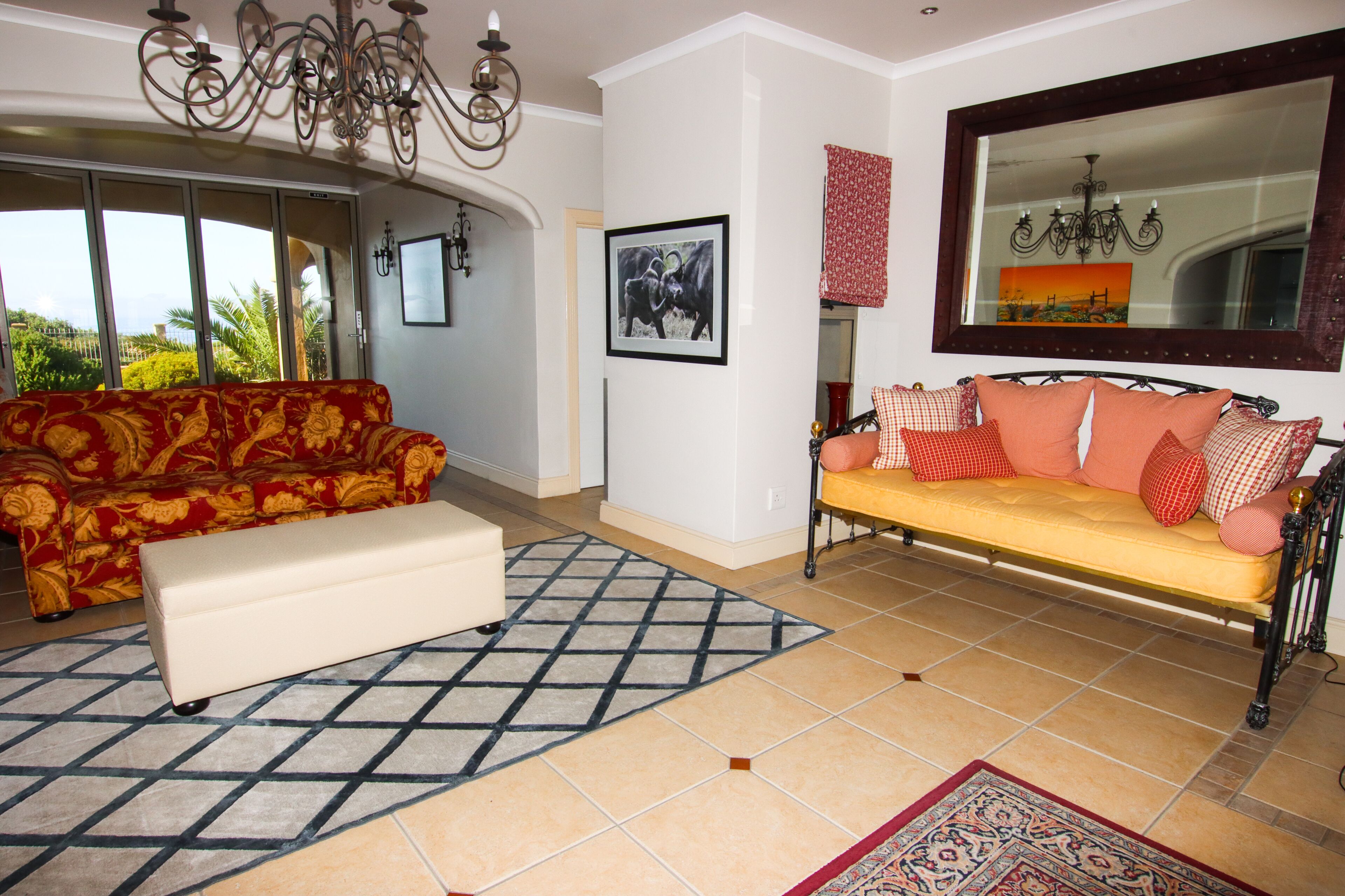 A living room with a sofa, ottoman, and a large mirror. There is a chandelier, a framed picture on the wall, and a window with a view of palm trees.