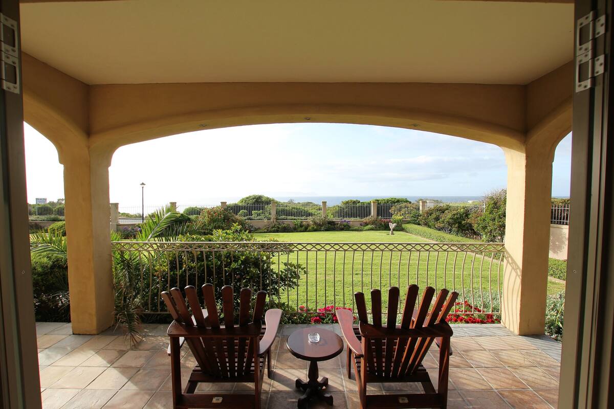 A covered patio with two wooden chairs and a small table, overlooking a well-maintained lawn and distant buildings.