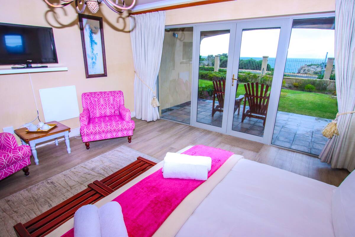 A hotel room with a bed, a TV, a chair, a small table, and a sliding glass door leading to a balcony with a view of the ocean.