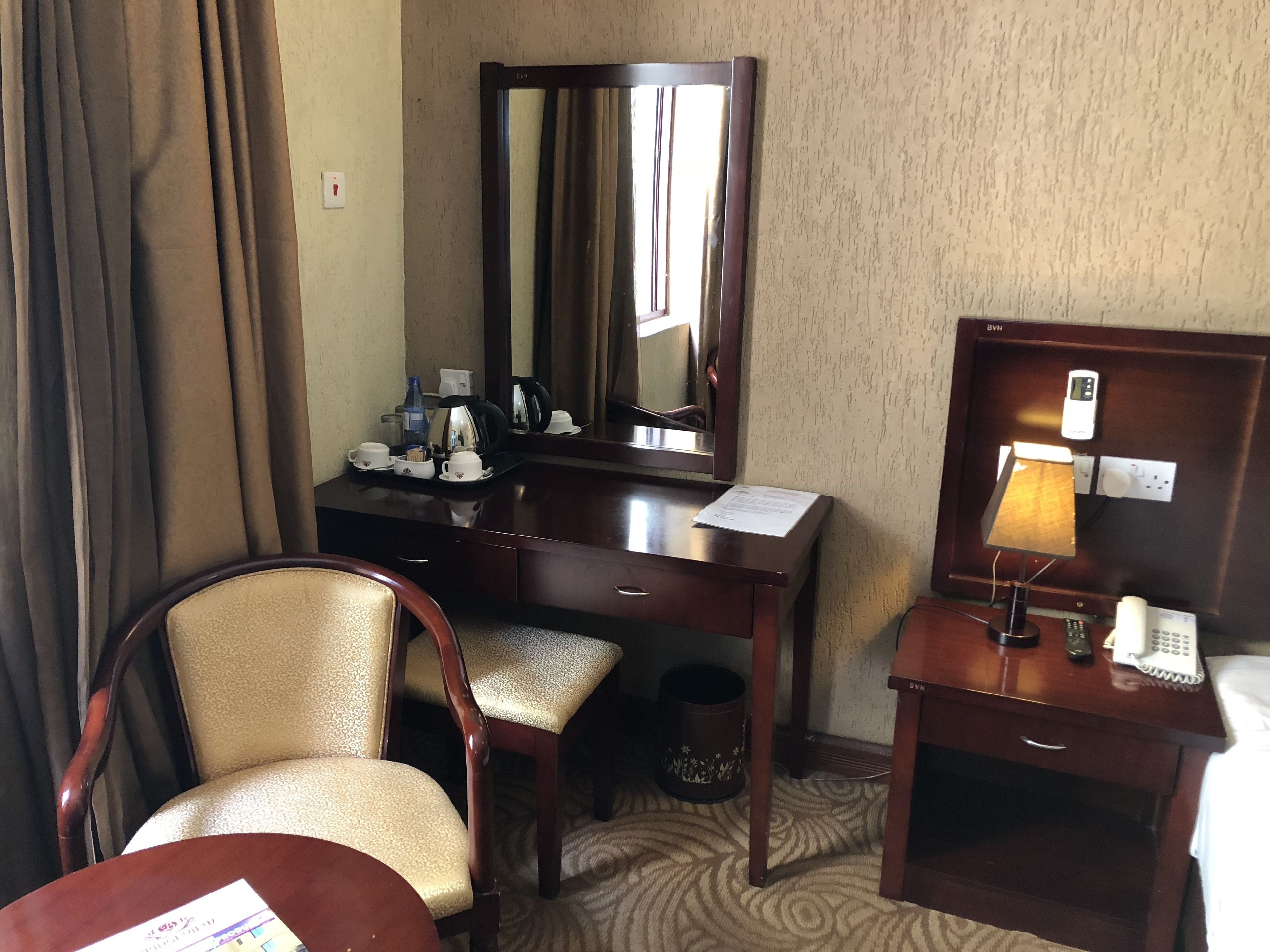 A hotel room with a wooden desk, a chair, a mirror, a lamp, a telephone, and a trash can.