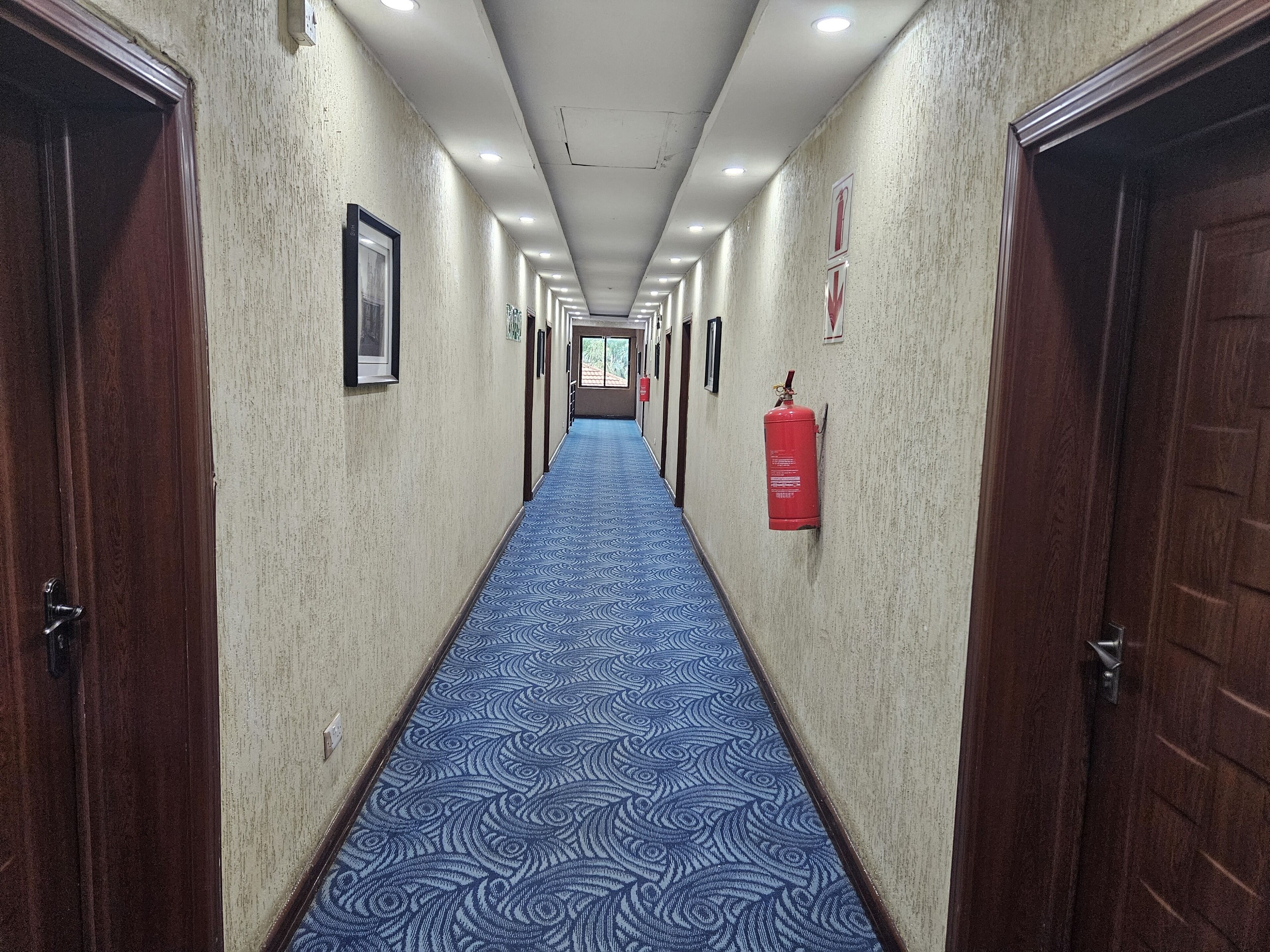 A long, narrow hallway with patterned carpet, wooden doors, and a fire extinguisher mounted on the wall.