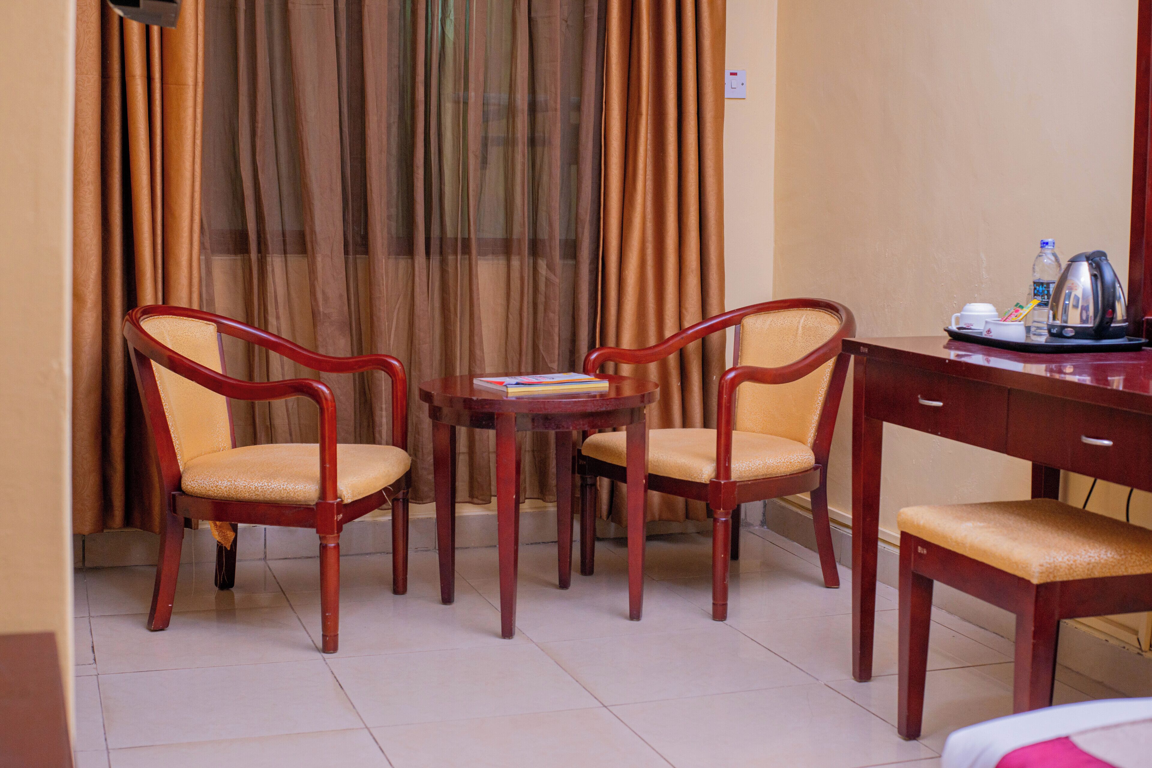 A hotel room with a wooden desk, two chairs, a small table, and a window with curtains.