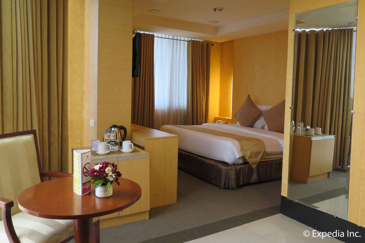 A hotel room with a bed, a desk with a telephone, a chair, a small table with a vase of flowers, and a mirror.