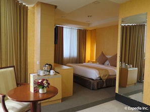 A hotel room with a bed, a desk with a telephone, a chair, a small table with a vase of flowers, and a mirror.