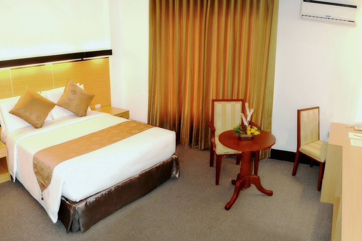 A hotel room with a large bed, a small round table with a vase, a chair, a desk with a computer, and a window with curtains.