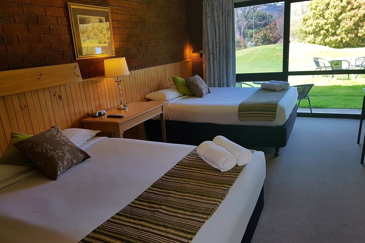 Habitación de hotel con dos camas, cabecera de madera, mesita de noche con lámpara y vista a un jardín a través de un ventanal grande.