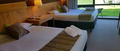 A hotel room with two beds, a wooden headboard, a bedside table with a lamp, and a view of a garden through a large window.