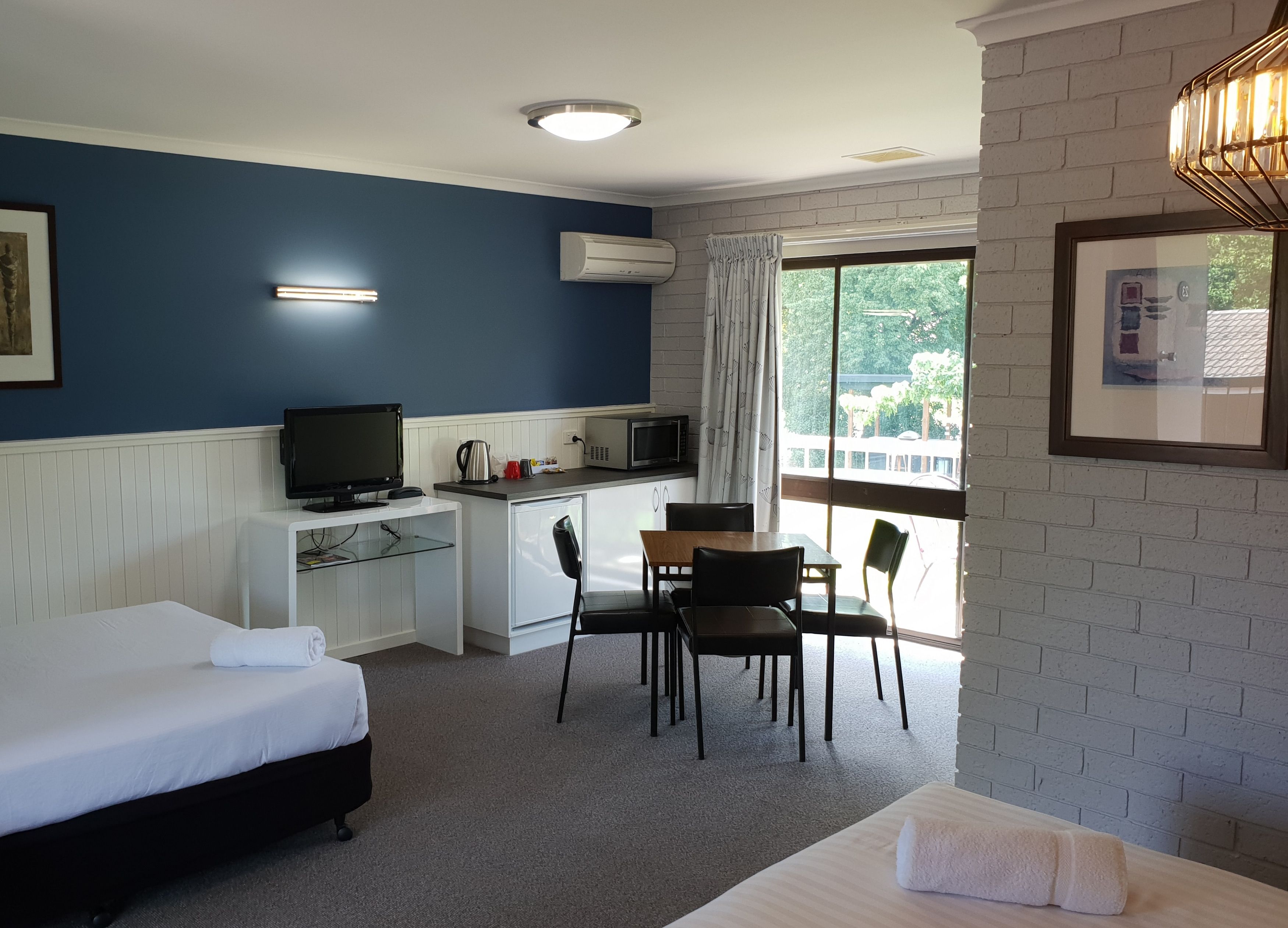 A hotel room with a bed, a desk with a computer, a small kitchenette, and a large window offering a view of greenery.
