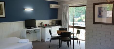 A hotel room with a bed, a desk with a computer, a small kitchenette, and a large window offering a view of greenery.