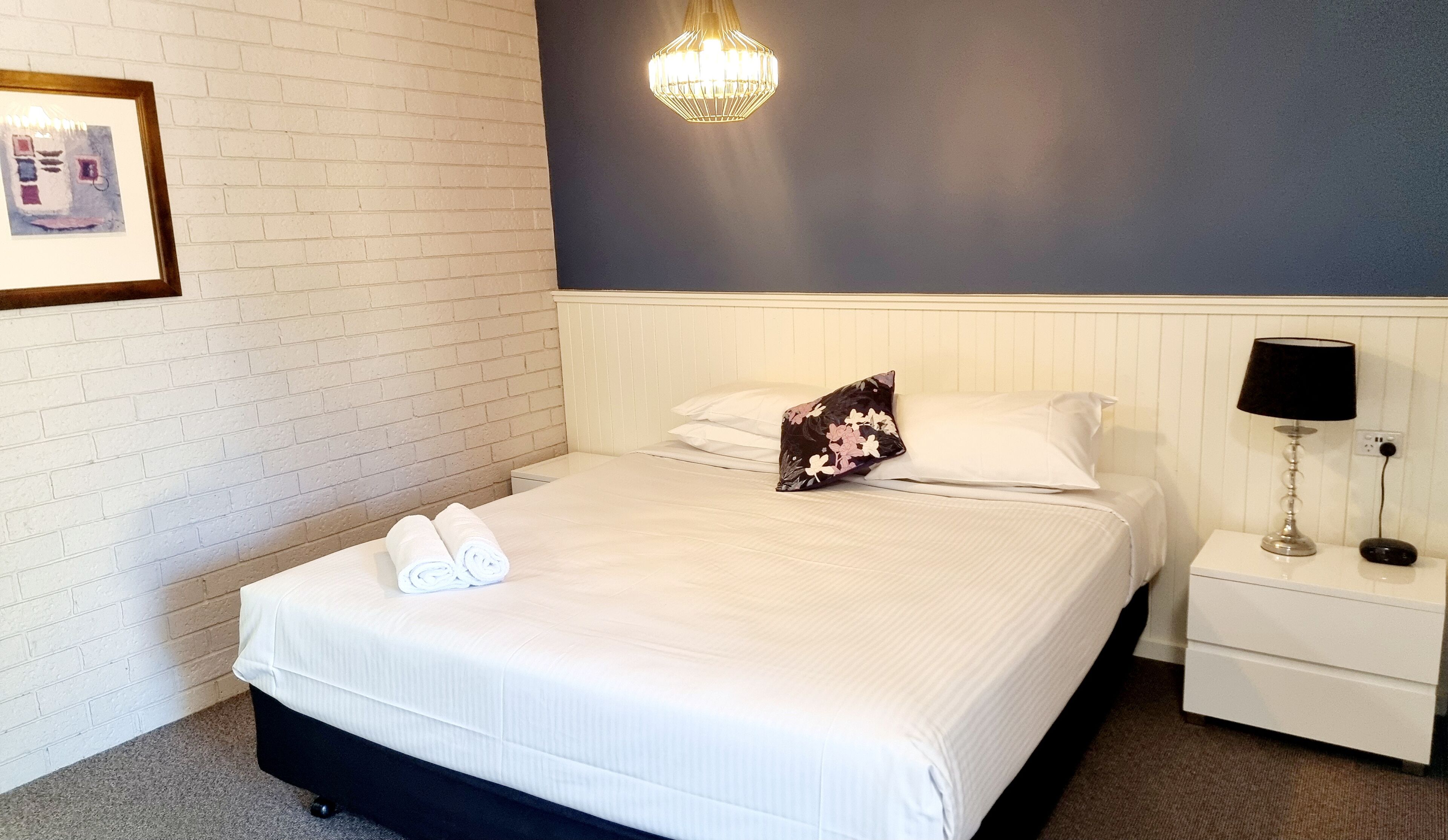 A neatly made bed with white linens and a floral pillow, a bedside lamp, and a framed picture on the wall.