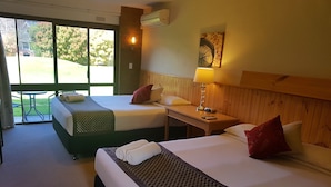 A hotel room with two beds, a wooden headboard, a table with a lamp, and a view of a garden through the window. - Motel Sierra (Bright)