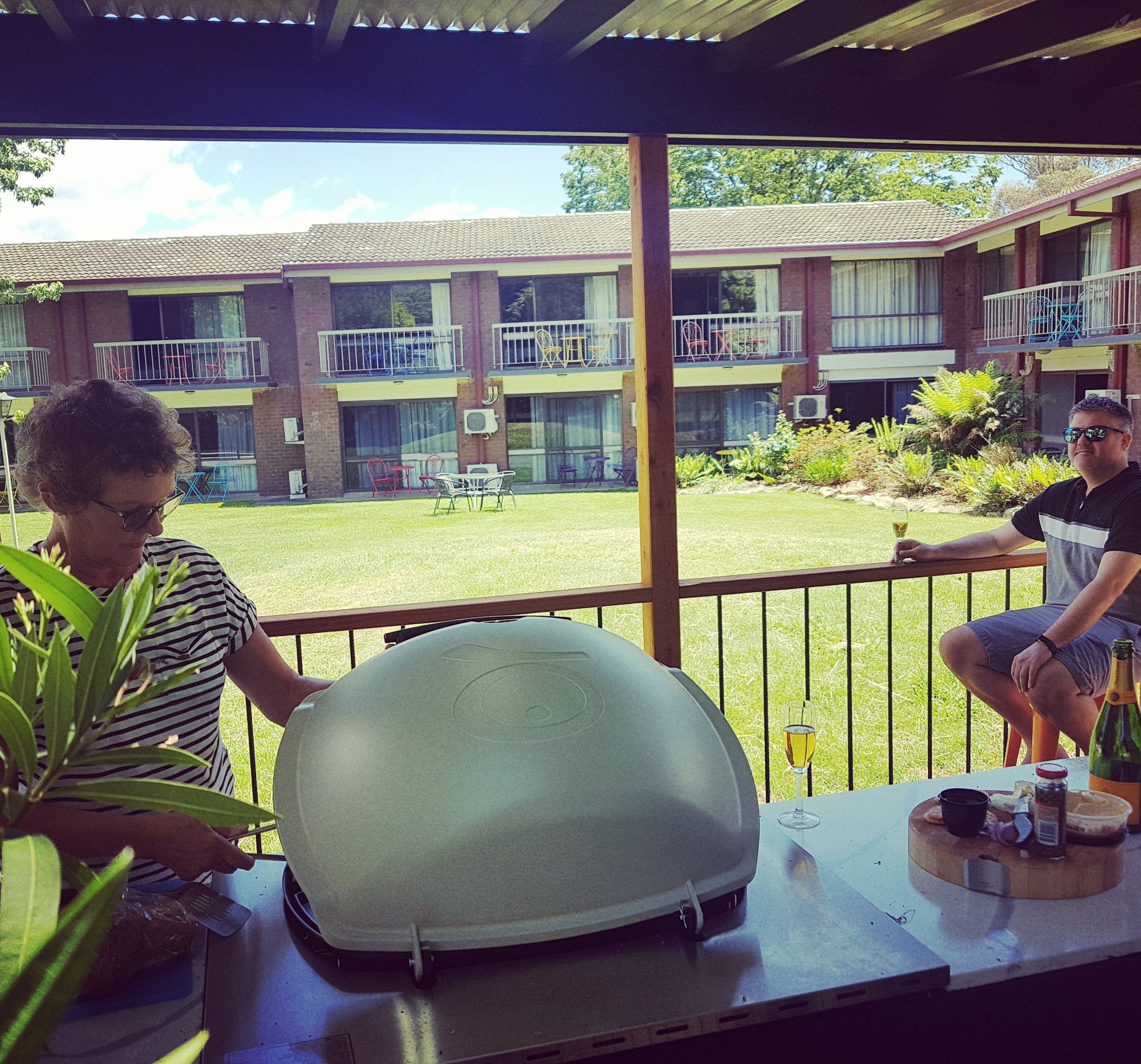 A covered outdoor area with a grill, a table with a bottle and glass, and a view of a building with balconies and a lawn.